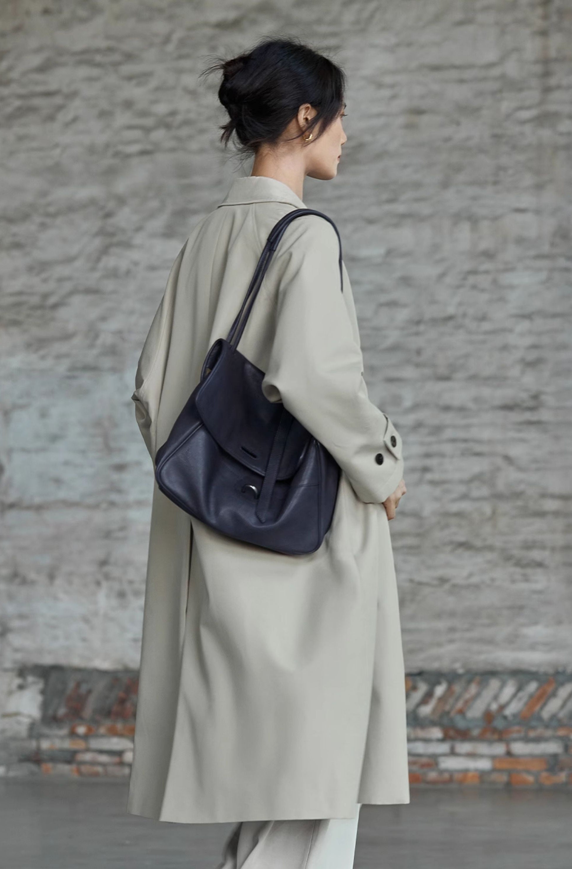 Person wearing a long coat with a navy blue bag against a textured wall.