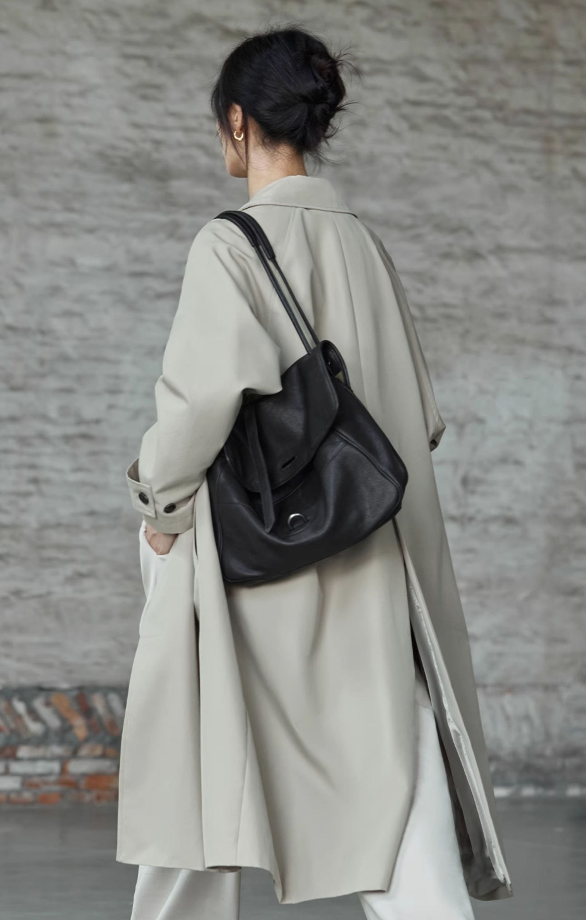 Person wearing a long beige coat and carrying a black backpack against a textured wall.
